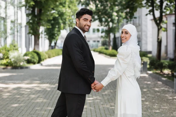 Novio feliz cogido de la mano con la novia musulmana sonriente en vestido de novia y caminando juntos - foto de stock