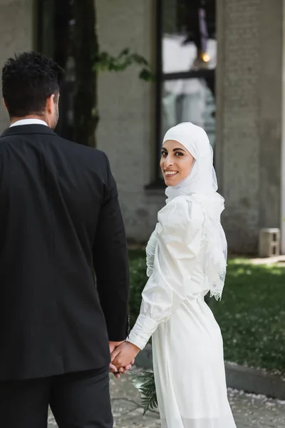 Groom holding hands with cheerful muslim bride in wedding dress and walking together — Stock Photo