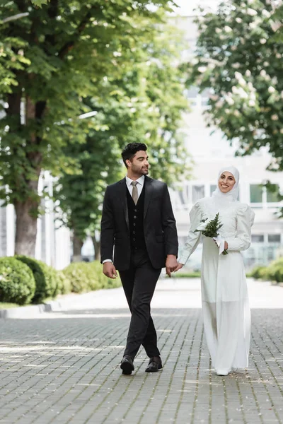 Счастливый жених держит за руки улыбающуюся мусульманскую невесту в хиджабе со свадебным букетом — стоковое фото