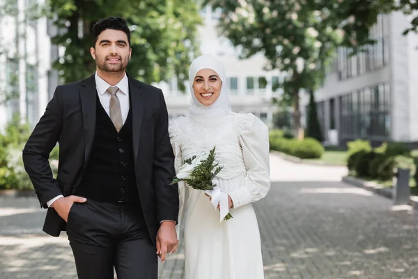 Retrato de novio feliz cogido de la mano con la novia musulmana sonriente en hijab con ramo de boda - foto de stock