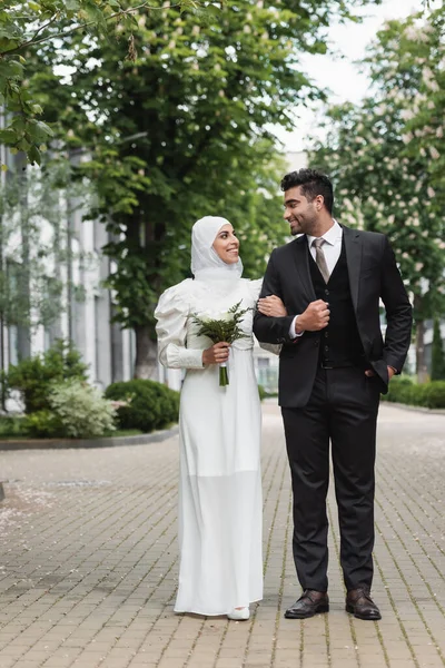 Pleine longueur de mariée musulmane heureuse en hijab de mariage et robe blanche tenant bouquet près du marié à l'extérieur — Photo de stock