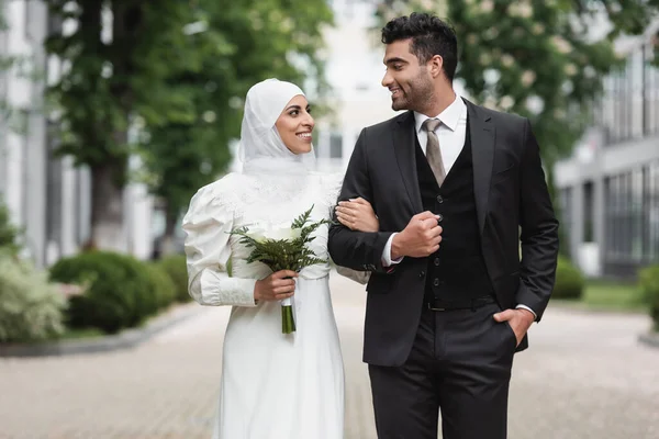 Glückliche muslimische Braut im Hochzeits-Hijab und weißem Kleid mit Blumenstrauß in der Nähe des Bräutigams draußen — Stockfoto