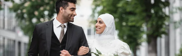 Happy muslim bride in wedding hijab and white dress holding bouquet near groom outside, banner — Stock Photo