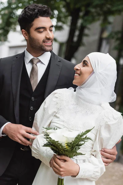 Alegre novia musulmana en hijab boda y vestido blanco celebración de ramo y mirando al novio fuera - foto de stock