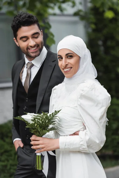 Fröhlicher Bräutigam blickt muslimische Braut im Hochzeits-Hijab und weißem Kleid mit Blumenstrauß an — Stockfoto