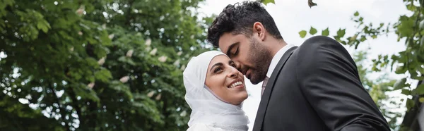 Novio en traje abrazando alegre novia musulmana en hijab blanco, bandera - foto de stock