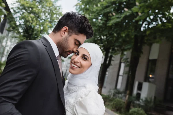 Novio en traje abrazando alegre novia musulmana en hijab y vestido blanco - foto de stock