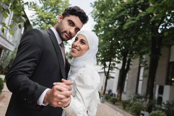 Feliz musulmán recién casados tomados de la mano y sonriendo en el parque verde - foto de stock