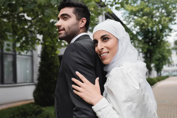 Noiva muçulmano satisfeito em hijab e vestido de noiva abraçando noivo — Fotografia de Stock