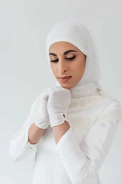 Retrato de la novia musulmana en hijab blanco y vestido de novia mirando hacia abajo aislado en gris - foto de stock