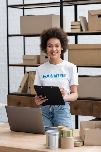 Mujer afroamericana feliz de pie con portapapeles cerca de la computadora portátil en el centro de voluntarios - foto de stock