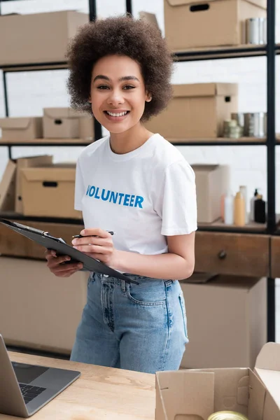 Happy african american woman with clipboard and pen smiling at camera in volunteer warehouse — Stock Photo