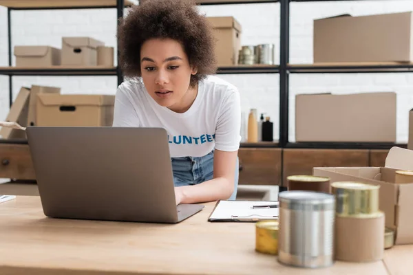 Afrikanisch-amerikanischer Freiwilliger arbeitet an Laptop in der Nähe verschwommener Dosen in Wohltätigkeitszentrum — Stockfoto