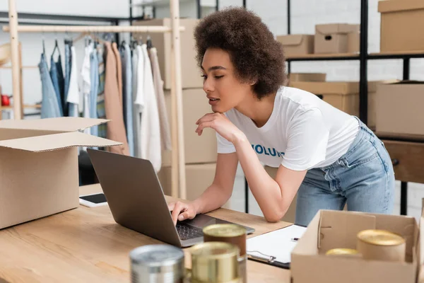 Young african american woman working on laptop near canned food on blurred foreground — Stock Photo