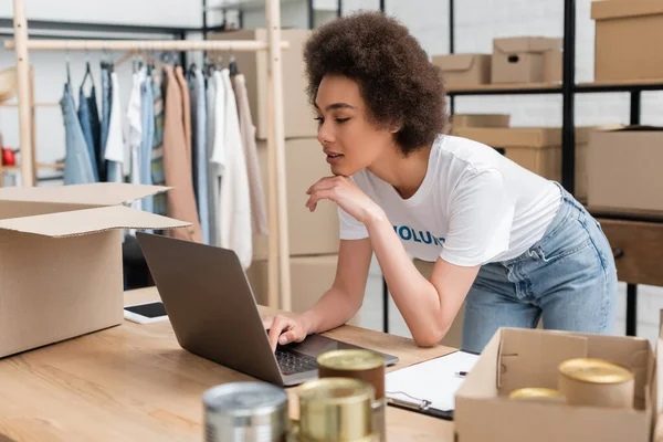 Jovem afro-americana trabalhando no laptop perto de comida enlatada em primeiro plano — Fotografia de Stock