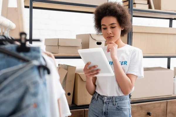 Volontaire afro-américain coûteux regardant tablette numérique près de rack avec boîtes en carton — Photo de stock