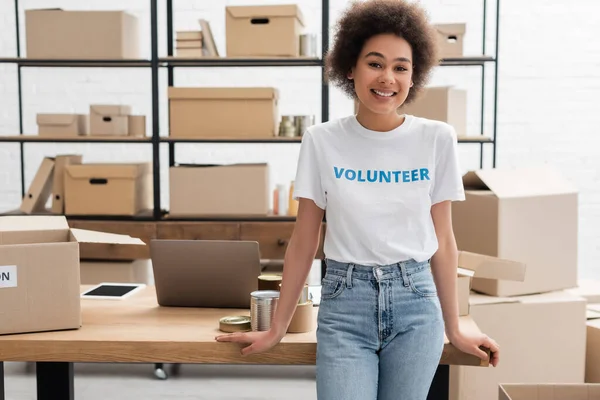 Junge afrikanisch-amerikanische Freiwillige lächelt in die Kamera neben Schreibtisch mit Gadgets in Charity-Zentrum — Stockfoto
