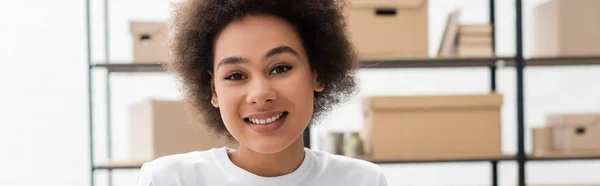 Cheerful african american volunteer smiling at camera in donation warehouse, banner — Stockfoto