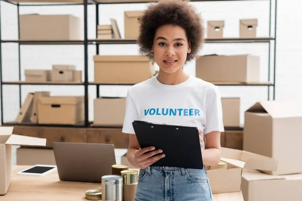 Young african american woman with clipboard looking at camera in volunteer warehouse — Stock Photo