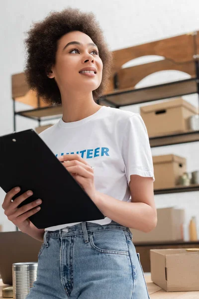 Low angle view of smiling african american volunteer with clipboard looking away in donation center — Fotografia de Stock