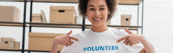 Heureuse femme afro-américaine pointant du doigt le lettrage bénévole sur t-shirt, bannière — Photo de stock