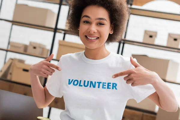 Mujer afroamericana feliz señalando letras de voluntarios en la camiseta en el centro de caridad - foto de stock