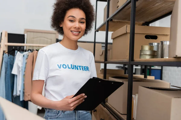 Happy african american woman with clipboard near rack with boxes and canned food — Photo de stock