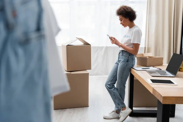 Full length of african american volunteer with smartphone near work desk and boxes in charity center - foto de stock