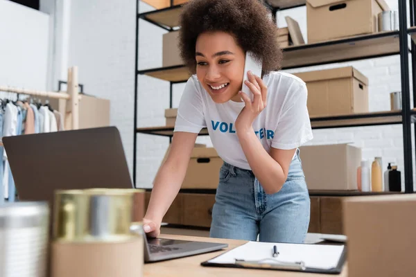 Happy african american volunteer talking on smartphone near laptop in donation center — Stock Photo