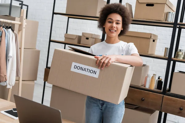 Young african american volunteer holding box with donation lettering in warehouse — Stockfoto