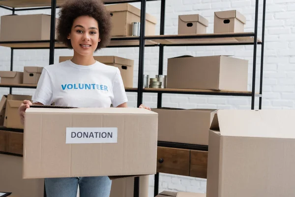 Volontario afroamericano sorridente alla macchina fotografica mentre tiene la scatola delle donazioni nel magazzino di beneficenza — Foto stock