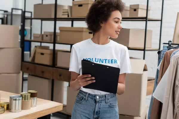 African american volunteer with clipboard looking at garments in charity center — Photo de stock