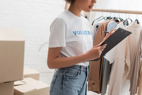 Cropped view of african american volunteer writing on clipboard near blurred garments - foto de stock