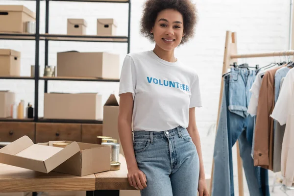 Joven afroamericano voluntario sonriendo en cámara cerca de alimentos enlatados y prendas borrosas - foto de stock