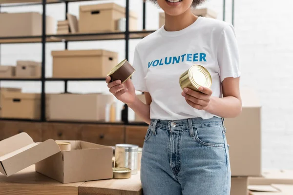 Partial view of african american volunteer holding canned food in charity storehouse — Stock Photo