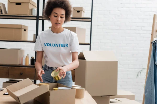 African american volunteer packing canned foodstuff in donation storehouse - foto de stock