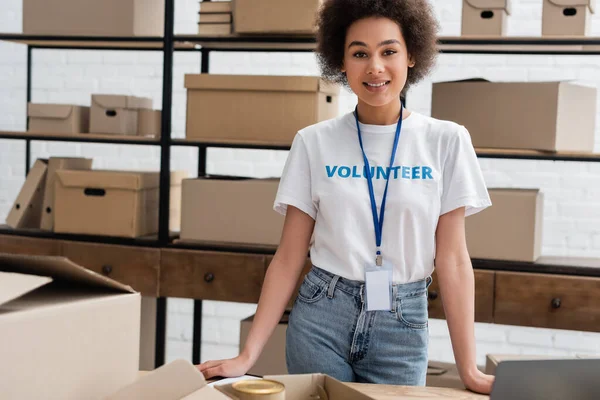 Young african american volunteer with name tag smiling at camera near carton boxes — Fotografia de Stock