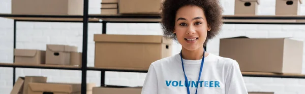 Молодий афроамериканський волонтер посміхається на камеру біля розмитої стійки з коробками, банер — стокове фото