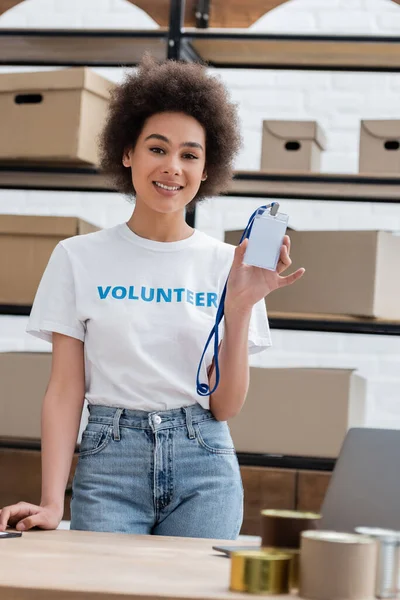 Happy african american volunteer showing blank name tag at camera in charity center — Stock Photo