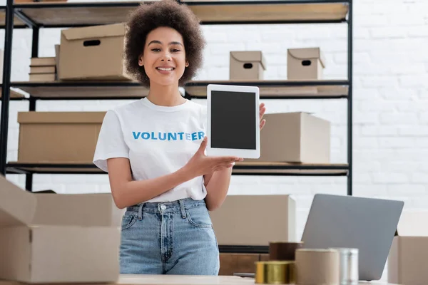 Heureux bénévole afro-américain montrant tablette numérique avec écran blanc dans l'entrepôt de don — Photo de stock