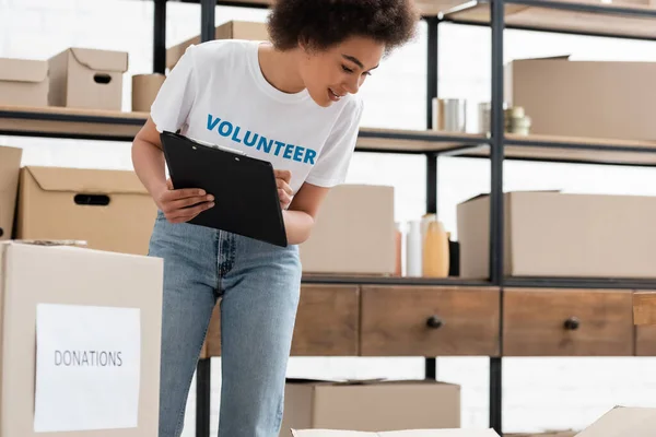 Young african american woman with clipboard looking at carton boxes in donation warehouse — Fotografia de Stock