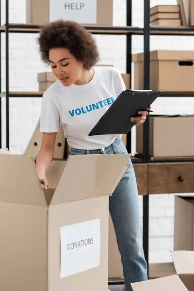Young african american volunteer with clipboard checking carton box in donation warehouse — Foto stock