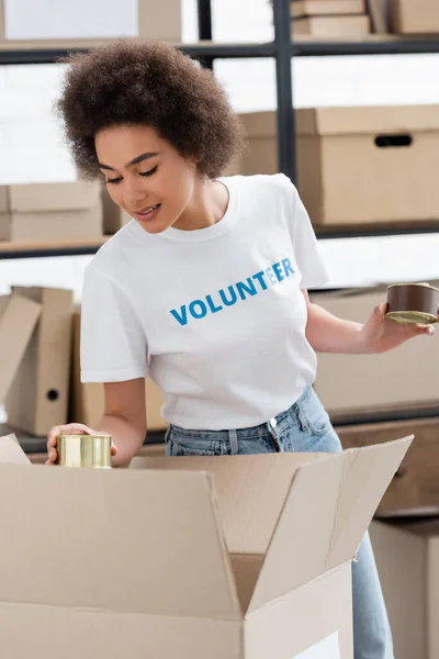 Smiling african american volunteer holding canned food near box in charity storehouse — Photo de stock