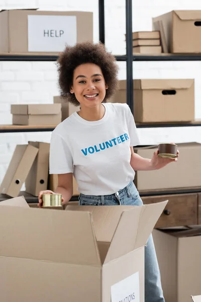 Joyful african american volunteer packing canned foodstuff in cardboard box — Stockfoto