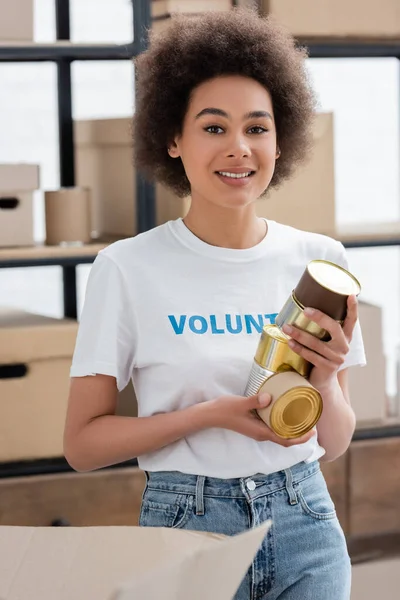 Young african american woman with canned food smiling at camera in volunteer center — Stock Photo