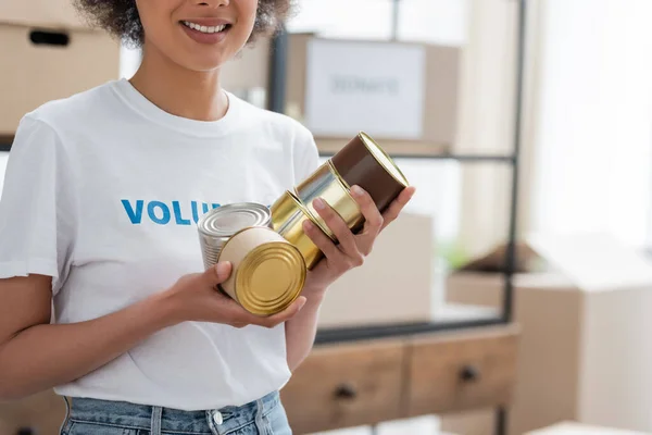 Cropped view of smiling african american volunteer holding canned food in charity warehouse — Stock Photo