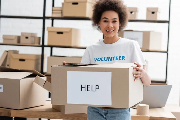 Feliz Africano americano voluntário segurando caixa de papelão com ajuda lettering no centro de caridade — Fotografia de Stock