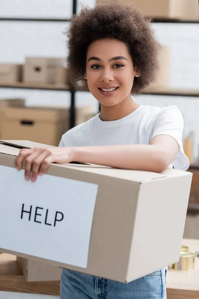 Jovem voluntário afro-americano sorrindo para a câmera perto da caixa com ajuda lettering — Fotografia de Stock