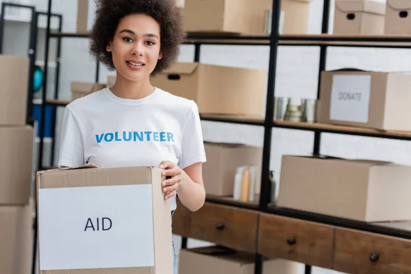 Smiling african american volunteer holding box with aid lettering in charity storehouse — Fotografia de Stock