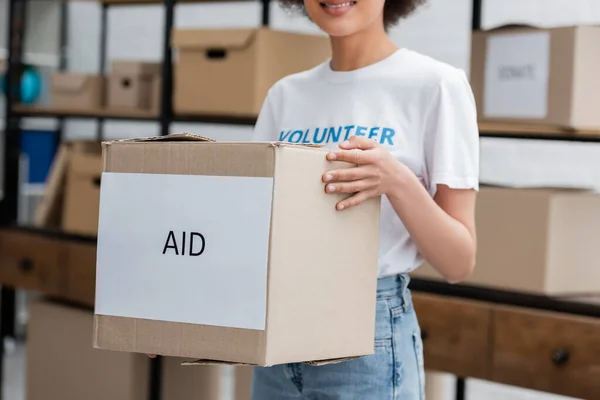 Cropped view of african american volunteer holding box with aid lettering in charity warehouse — Fotografia de Stock